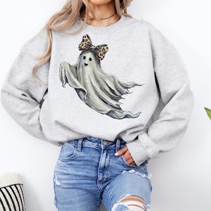 May include: A light grey sweatshirt with a playful ghost design. The ghost is depicted with a flowing white form, a smiling face, and a leopard print bow. Ideal for Halloween or anyone who enjoys fun, spooky clothing.