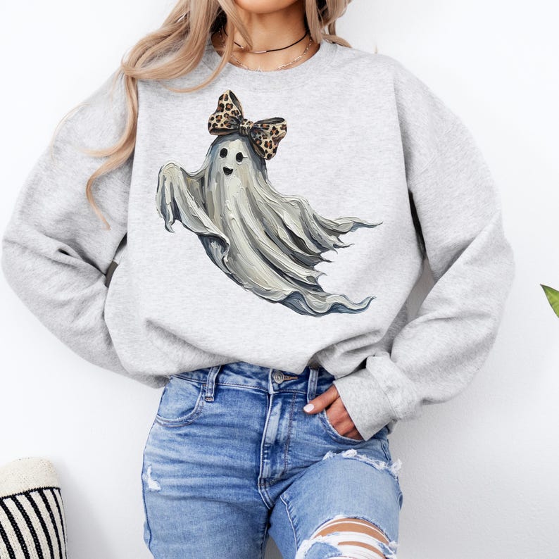 May include: A light grey sweatshirt with a playful ghost design. The ghost is depicted with a flowing white form, a smiling face, and a leopard print bow. Ideal for Halloween or anyone who enjoys fun, spooky clothing.