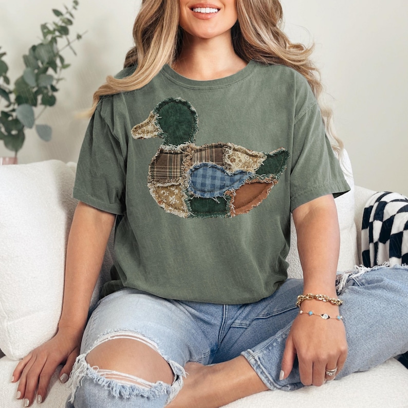 May include: An olive green short-sleeve t-shirt featuring a large, central duck graphic composed of various fabric patches. The duck design incorporates multiple textures and patterns, including plaid, floral, and solid brown and blue fabric pieces.