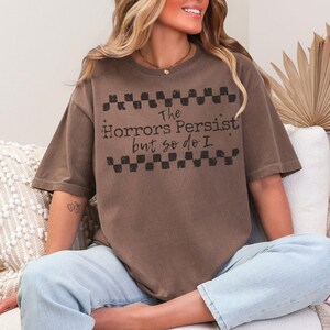 May include: Brown t-shirt with the text "The Horrors Persist but so do I" in black. The shirt has a checkerboard design above and below the text. The shirt is a relaxed fit and has short sleeves.