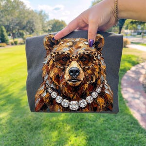 May include: A dark gray t-shirt featuring a detailed, artistic rendering of a bear's face. The bear wears leopard print glasses and a diamond necklace. The artwork uses a palette of browns, golds, and whites.