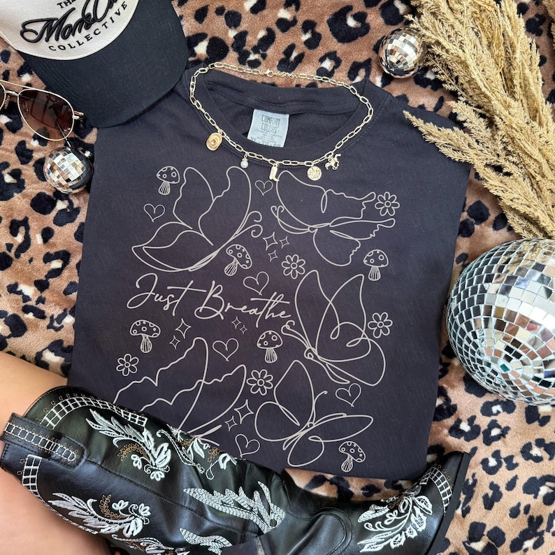 Puede incluir: Una camiseta negra con un dise&ntilde;o de mariposas y setas blancas, y el texto "Just Breathe". La imagen tambi&eacute;n incluye una gorra negra, gafas de sol, un collar dorado y botas de vaquero negras.