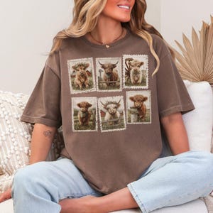 May include: Brown t-shirt featuring six postage stamp-style images of Highland cows. Each image showcases a different cow in a pastoral setting. The shirt has a relaxed fit and short sleeves, suitable for casual wear.