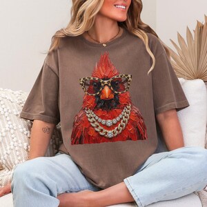 May include: Brown t-shirt featuring a vibrant red cardinal wearing leopard print glasses and a gold chain necklace. The bird has a detailed, textured appearance. The shirt is a relaxed fit, with short sleeves.