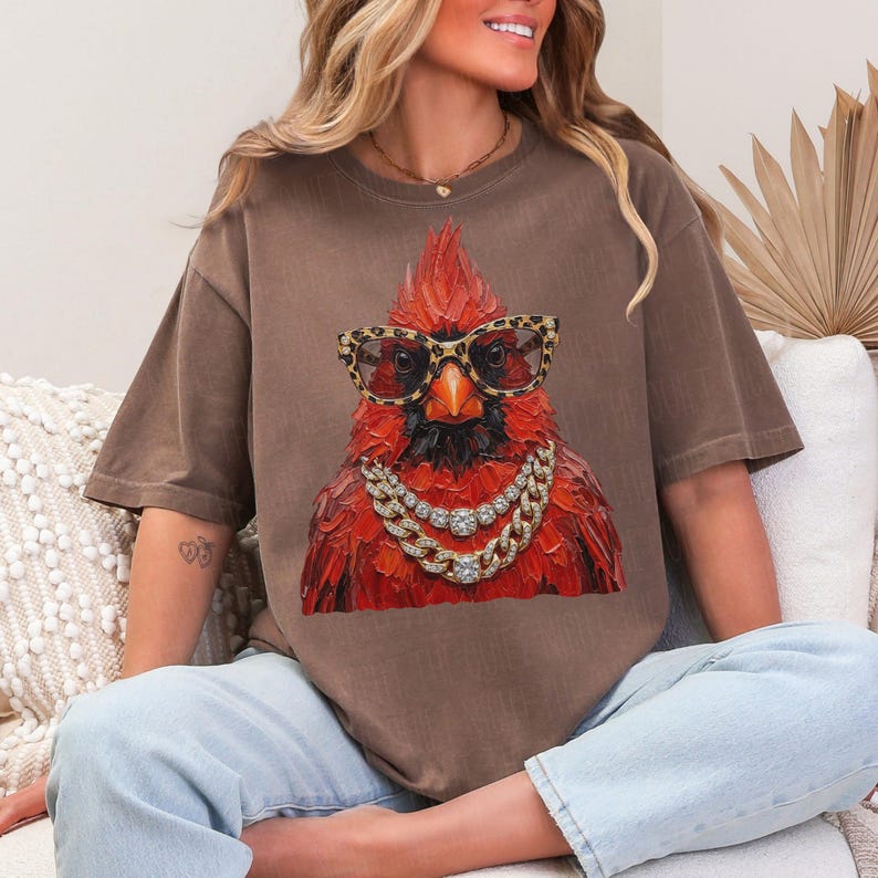 May include: Brown t-shirt featuring a vibrant red cardinal wearing leopard print glasses and a gold chain necklace. The bird has a detailed, textured appearance. The shirt is a relaxed fit, with short sleeves.