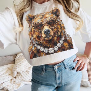 May include: White t-shirt featuring a detailed illustration of a bear wearing leopard print glasses and a diamond necklace. The bear's fur is rendered in shades of brown, gold, and cream. The shirt is a casual, everyday wear item.