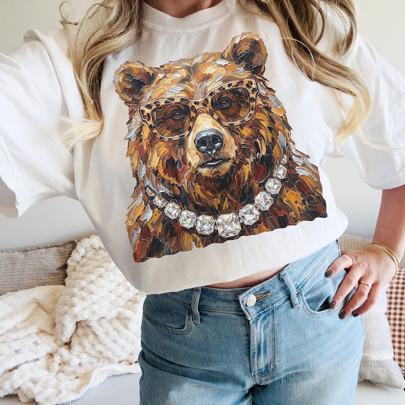May include: White t-shirt featuring a detailed illustration of a bear wearing leopard print glasses and a diamond necklace. The bear's fur is rendered in shades of brown, gold, and cream. The shirt is a casual, everyday wear item.