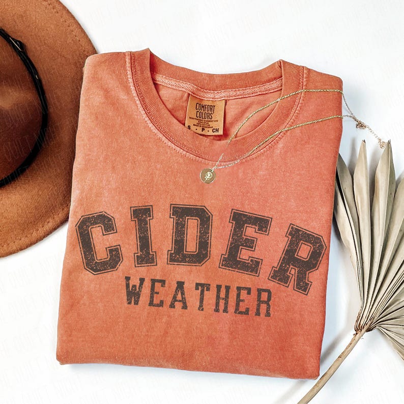 K&ouml;nnte beinhalten: Ein orange-rotes T-Shirt mit den Worten "CIDER WEATHER" in dunkelbraunen Blockbuchstaben. Das Shirt hat einen Rundhalsausschnitt und ein kleines Etikett am Ausschnitt. Eine goldene Halskette und ein brauner Hut sind ebenfalls im Bild.