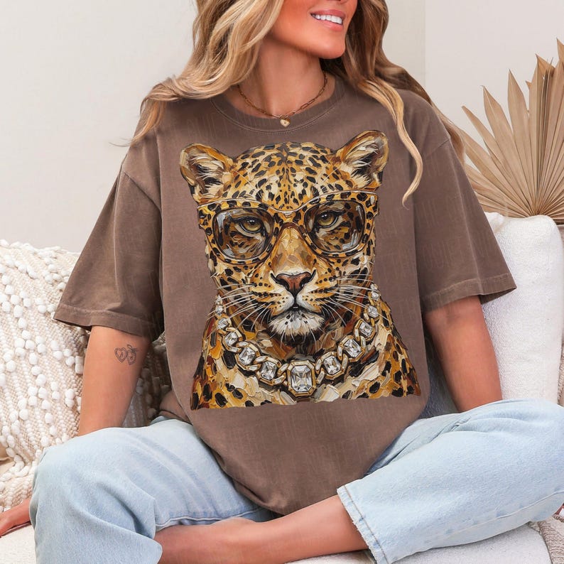 Puede incluir: Camiseta marr&oacute;n con una ilustraci&oacute;n detallada de un leopardo con gafas y un collar de diamantes. El pelaje del leopardo tiene manchas marrones y beige. Las gafas tienen montura dorada y el collar tiene grandes gemas cuadradas.