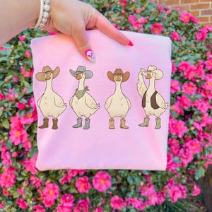 May include: A light pink sweatshirt featuring a cartoon design of four geese dressed as cowboys. Each goose wears a cowboy hat and boots, with one also sporting a vest and another a bandana. The design is set against a background of pink flowers.