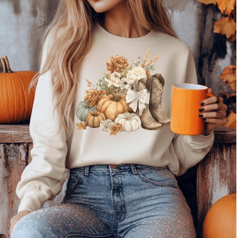 May include: Cream-colored sweatshirt featuring a fall-themed graphic. The design includes pumpkins, flowers, and cowboy boots. An orange mug is held in the hand. The person is wearing blue jeans.