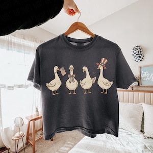 May include: Dark gray t-shirt featuring a whimsical illustration of four geese. Each goose is adorned with patriotic accessories, including an American flag, a bow, and a top hat. The design is printed on a comfortable, casual tee.