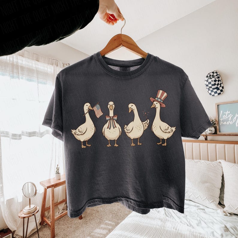 May include: Dark gray t-shirt featuring a whimsical illustration of four geese. Each goose is adorned with patriotic accessories, including an American flag, a bow, and a top hat. The design is printed on a comfortable, casual tee.