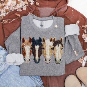 Peut inclure: Un sweat-shirt gris avec des t&ecirc;tes de chevaux brod&eacute;es en marron, noir et cr&egrave;me. Le sweat-shirt fait partie d'une tenue d&eacute;contract&eacute;e, avec un sweat &agrave; capuche marron et un jean bleu en arri&egrave;re-plan. Un collier dor&eacute; et un chouchou blanc sont &eacute;galement pr&eacute;sents.