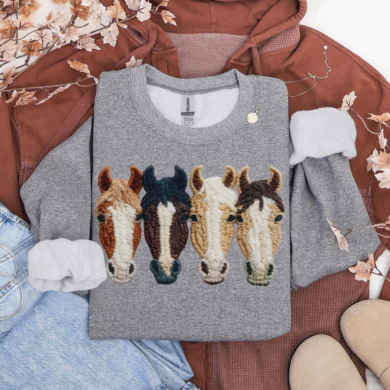 Peut inclure: Un sweat-shirt gris avec des t&ecirc;tes de chevaux brod&eacute;es en marron, noir et cr&egrave;me. Le sweat-shirt fait partie d'une tenue d&eacute;contract&eacute;e, avec un sweat &agrave; capuche marron et un jean bleu en arri&egrave;re-plan. Un collier dor&eacute; et un chouchou blanc sont &eacute;galement pr&eacute;sents.