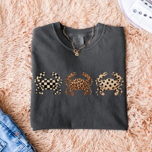 以下が含まれることがあります： 3匹のカニのデザインが施された濃いグレーのTシャツ。カニは、白黒の市松模様、ヒョウ柄、黒い水玉模様のタンなど、ユニークなパターンを持っています。ゴールドのチェーンネックレスが首の近くに見えます。
