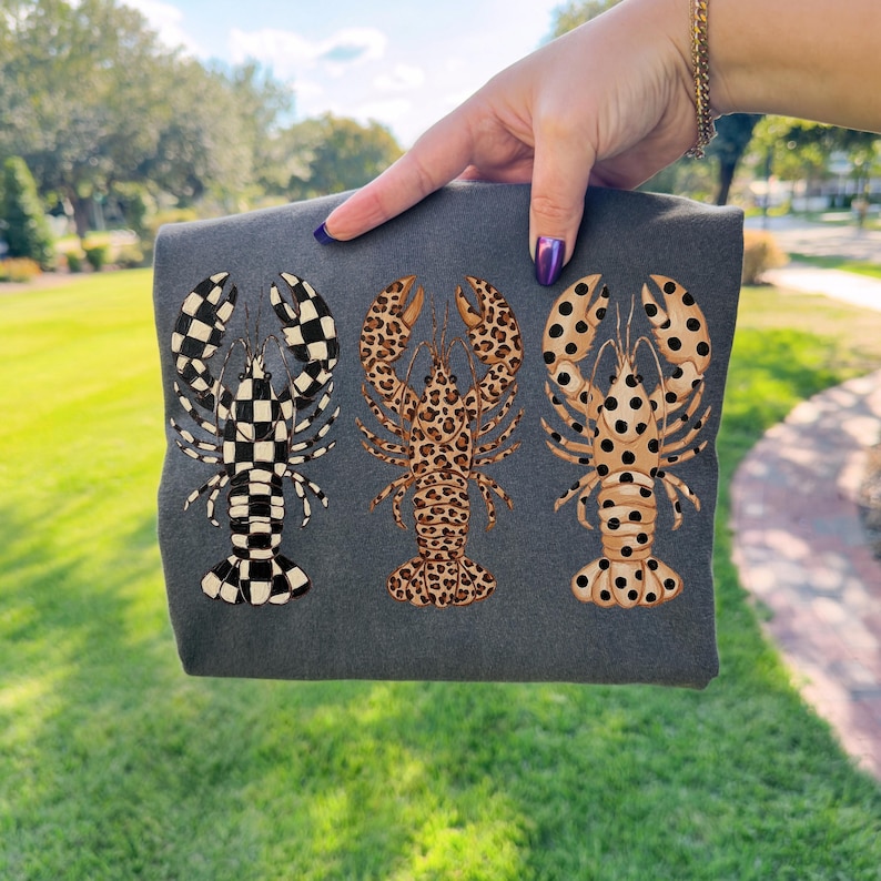 May include: Dark gray folded t-shirt featuring three lobster designs. The lobsters have unique patterns: a black and white checkerboard, a leopard print, and a tan with black polka dots. The shirt is held up by a person.
