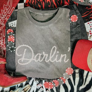 Pu&ograve; includere: Una maglietta grigia con la scritta "Darlin'" in un carattere perlato. La maglietta &egrave; abbinata a pantaloncini di jeans, un berretto da baseball rosso e stivali da cowboy rossi. Gettoni da poker e dadi sono sparsi intorno agli articoli.
