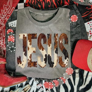 K&ouml;nnte beinhalten: Ein graues Rundhals-T-Shirt mit dem Wort "JESUS" im Kuhfellmuster. Das Bild zeigt Jeansshorts, eine rote Kappe, rote Cowboystiefel, Pokerkarten, W&uuml;rfel und Spielkarten.