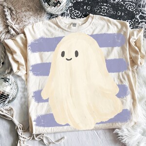 May include: A cream-colored short-sleeve t-shirt featuring a central graphic of a pale yellow ghost with a simple smiling face. The ghost is layered over a background of four horizontal, distressed-style light purple brushstroke stripes.
