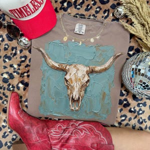 May include: A taupe Comfort Colors t-shirt with a textured bull skull painting on a teal background. The shirt is styled with a red baseball cap, sunglasses, a gold necklace, and red cowboy boots. A disco ball and leopard print fabric are also visible.