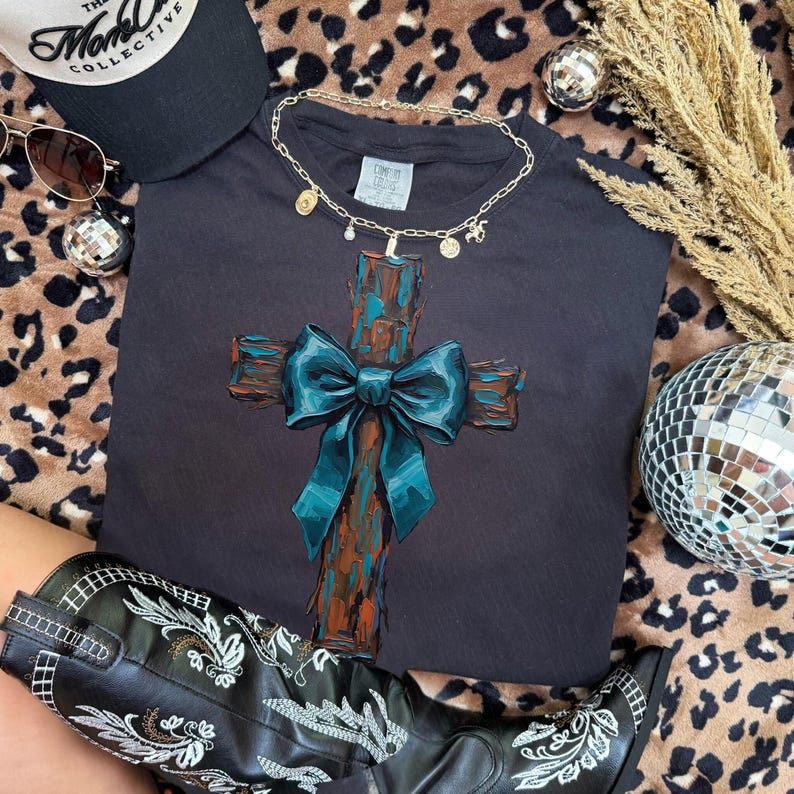 Peut inclure: Un t-shirt noir avec un motif de croix et un n&oelig;ud bleu sarcelle. La croix a un aspect textur&eacute; et rustique avec de la peinture marron et bleu sarcelle. Un collier dor&eacute; et un chapeau noir sont &eacute;galement visibles.