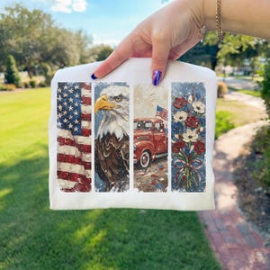 Puede incluir: Camiseta blanca con ilustraciones patri&oacute;ticas. El dise&ntilde;o incluye una bandera estadounidense, un &aacute;guila calva, una camioneta roja vintage con una bandera y un ramo de flores rojas, blancas y azules. La obra de arte tiene una apariencia texturizada y pintada.