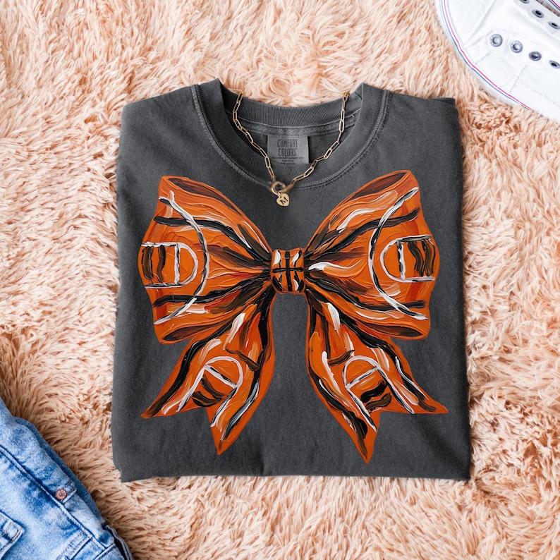 May include: Dark gray t-shirt featuring a large orange bow design with black and white accents. The bow has a basketball-like pattern. A gold necklace is visible. The shirt is on a pink fluffy surface.