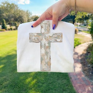 May include: A white sweatshirt featuring a textured cross design in shades of beige, gray, and brown. The cross has a painted, brushstroke appearance. The sweatshirt is held up against a background of green grass and trees.
