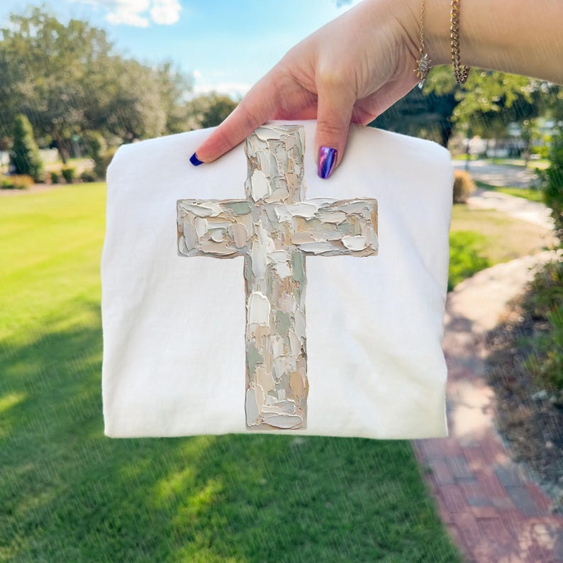 May include: A white sweatshirt featuring a textured cross design in shades of beige, gray, and brown. The cross has a painted, brushstroke appearance. The sweatshirt is held up against a background of green grass and trees.