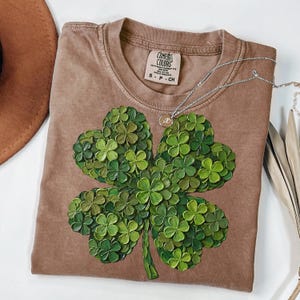 May include: A brown Comfort Colors t-shirt featuring a large, textured four-leaf clover design in shades of green. A silver necklace with a small pendant rests on the shirt. The shirt has a crew neck and short sleeves.