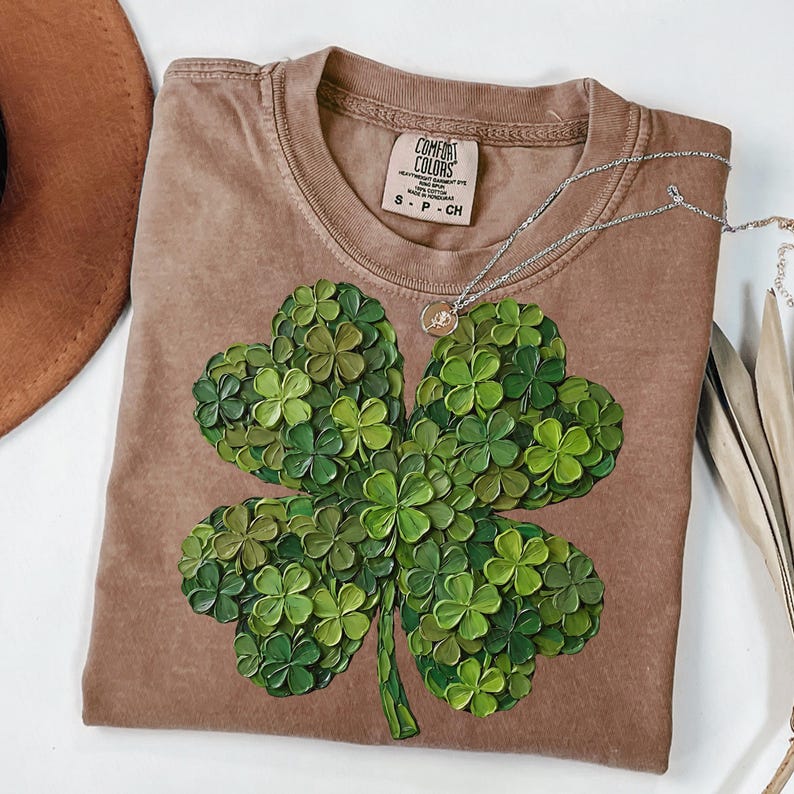 May include: A brown Comfort Colors t-shirt featuring a large, textured four-leaf clover design in shades of green. A silver necklace with a small pendant rests on the shirt. The shirt has a crew neck and short sleeves.