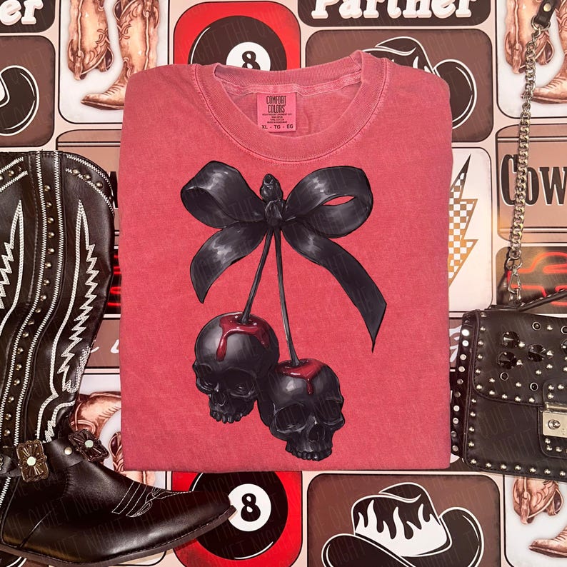 May include: Red t-shirt featuring a black bow and two skull-shaped cherries. The shirt has a crew neck and short sleeves. The tag reads "Comfort Colors". The shirt is folded on a patterned background with cowboy boots and a purse.