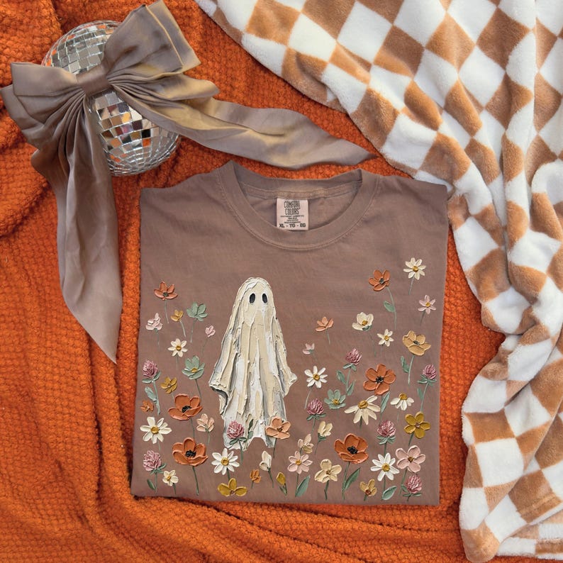 Może przedstawiać: Brązowy t-shirt z kr&oacute;tkim rękawem z centralną grafiką przedstawiającą białego ducha otoczonego polem teksturowanych, wielokolorowych kwiat&oacute;w w odcieniach pomarańczu, ż&oacute;łci, r&oacute;żu i bieli. Koszulka posiada okrągły dekolt oraz metkę.