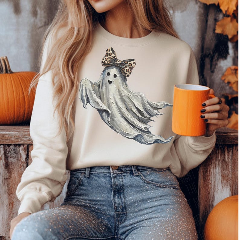 May include: A cream-coloured sweatshirt with a watercolour ghost wearing a leopard print bow. The ghost has a friendly expression. The person in the image is holding an orange mug. The sweatshirt is paired with blue jeans.