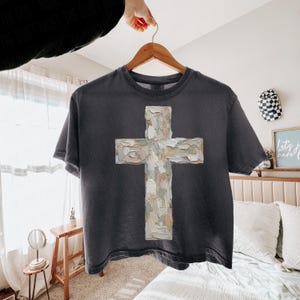 May include: Dark gray t-shirt featuring a textured cross design in shades of white, beige, and gray. The cross is centered on the shirt, which is hanging on a wooden hanger. The shirt has a vintage, worn look.