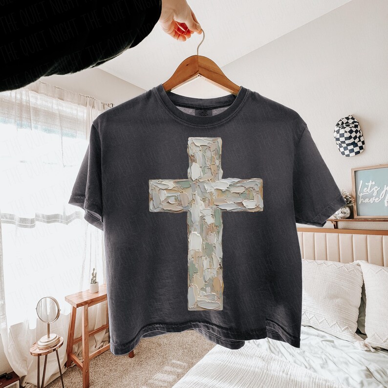 May include: Dark gray t-shirt featuring a textured cross design in shades of white, beige, and gray. The cross is centered on the shirt, which is hanging on a wooden hanger. The shirt has a vintage, worn look.