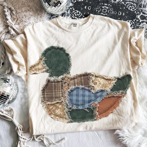 May include: A cream-colored short-sleeve t-shirt featuring a large, central duck-shaped appliqu&eacute;. The duck is constructed from various fabric patches, including green corduroy, brown plaid, blue gingham, and floral patterns with frayed edges.