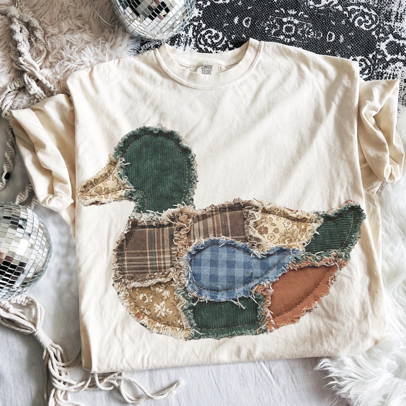 May include: A cream-colored short-sleeve t-shirt featuring a large, central duck-shaped appliqu&eacute;. The duck is constructed from various fabric patches, including green corduroy, brown plaid, blue gingham, and floral patterns with frayed edges.
