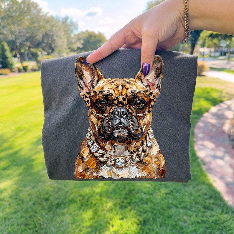May include: A dark gray t-shirt featuring a detailed illustration of a French Bulldog wearing leopard print glasses and a diamond-studded chain. The dog's fur is painted in shades of brown and tan.
