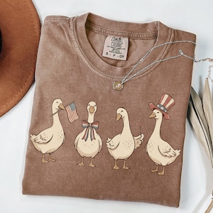 May include: Brown Comfort Colors t-shirt featuring a graphic of four cartoon geese. One goose holds an American flag, another wears a bow, and the last wears a patriotic hat. A silver necklace with a pendant is also visible.