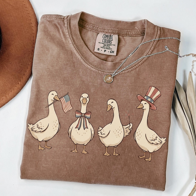 May include: Brown Comfort Colors t-shirt featuring a graphic of four cartoon geese. One goose holds an American flag, another wears a bow, and the last wears a patriotic hat. A silver necklace with a pendant is also visible.
