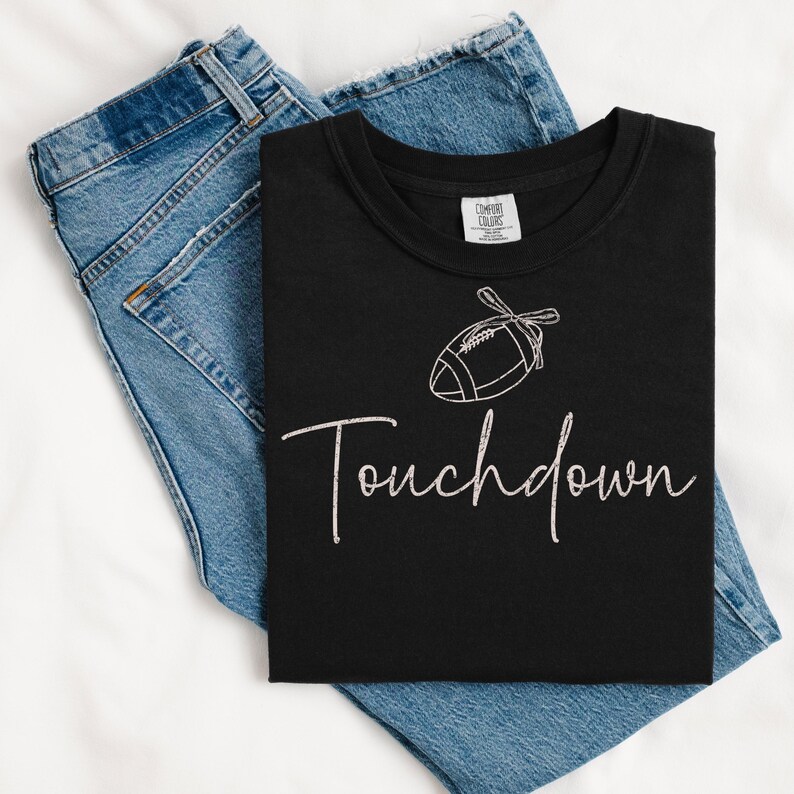 May include: Black t-shirt with a white football graphic and the word "Touchdown" in a script font. The shirt is folded on top of a pair of blue denim jeans.