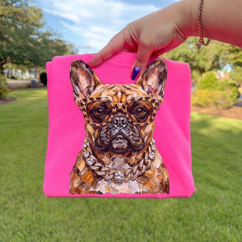 May include: A hot pink t-shirt featuring a detailed illustration of a French Bulldog wearing leopard print glasses and a diamond-studded chain. The dog's fur is a mix of brown and tan tones.