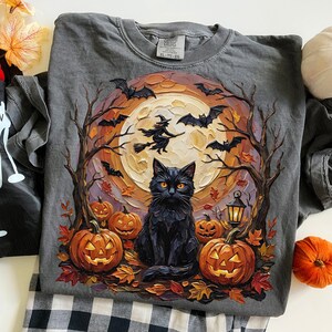 May include: A charcoal gray t-shirt featuring a graphic print of a black cat sitting in front of a large full moon. The design includes a witch on a broomstick, flying bats, bare trees, glowing jack-o'-lanterns, and scattered autumn leaves.