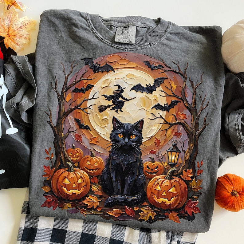 May include: A charcoal gray t-shirt featuring a graphic print of a black cat sitting in front of a large full moon. The design includes a witch on a broomstick, flying bats, bare trees, glowing jack-o'-lanterns, and scattered autumn leaves.