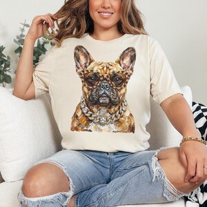 May include: A beige t-shirt featuring a watercolor painting of a French Bulldog wearing leopard print glasses and a diamond chain. The dog's fur is a mix of brown and tan. The shirt is worn by a person.