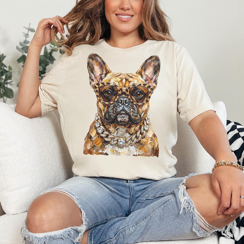 May include: A beige t-shirt featuring a watercolor painting of a French Bulldog wearing leopard print glasses and a diamond chain. The dog's fur is a mix of brown and tan. The shirt is worn by a person.