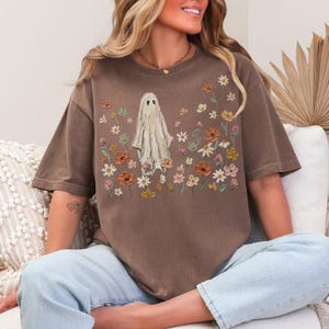 Może przedstawiać: Brązowy t-shirt z kr&oacute;tkim rękawem z centralną grafiką przedstawiającą białego ducha otoczonego r&oacute;żnorodnymi kolorowymi kwiatami w odcieniach pomarańczu, ż&oacute;łci i bieli. Wz&oacute;r na klatce piersiowej ma teksturę przypominającą malarstwo.