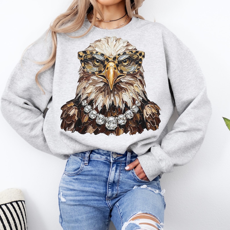 May include: A light gray sweatshirt featuring a detailed illustration of an eagle wearing leopard print glasses and a diamond necklace. The eagle's feathers are depicted in shades of brown and white. The sweatshirt is a casual, comfortable style.
